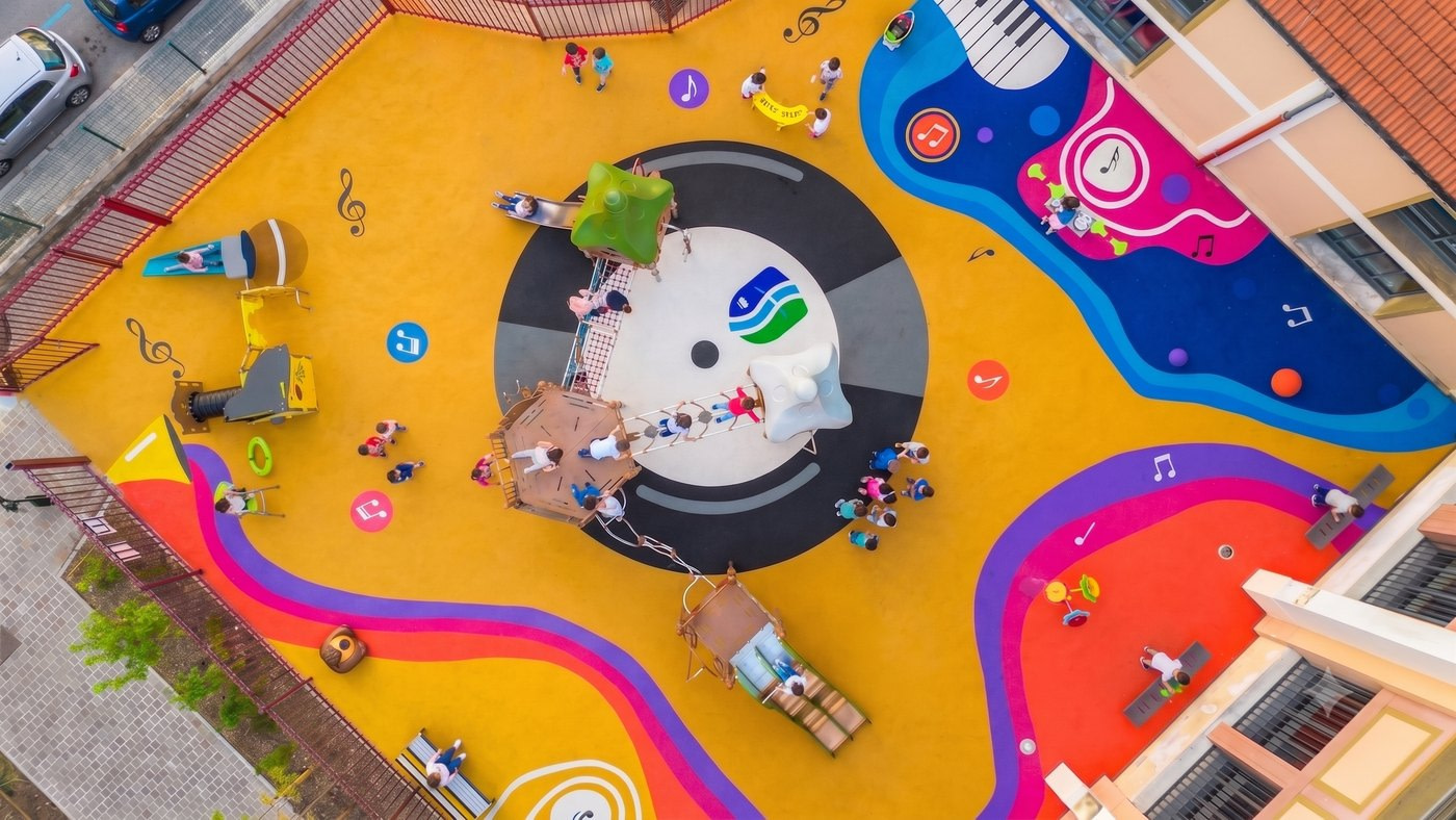 Cour d’école vue du dessus : sol souple jaune au motif de disque vinyle et de notes de musique, structures de jeux et zones colorées